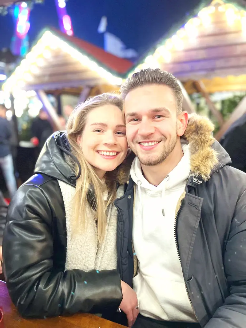 Zora und Kenny in Winterjacken, lachend unter den Lichterketten eines Weihnachtsmarkts
